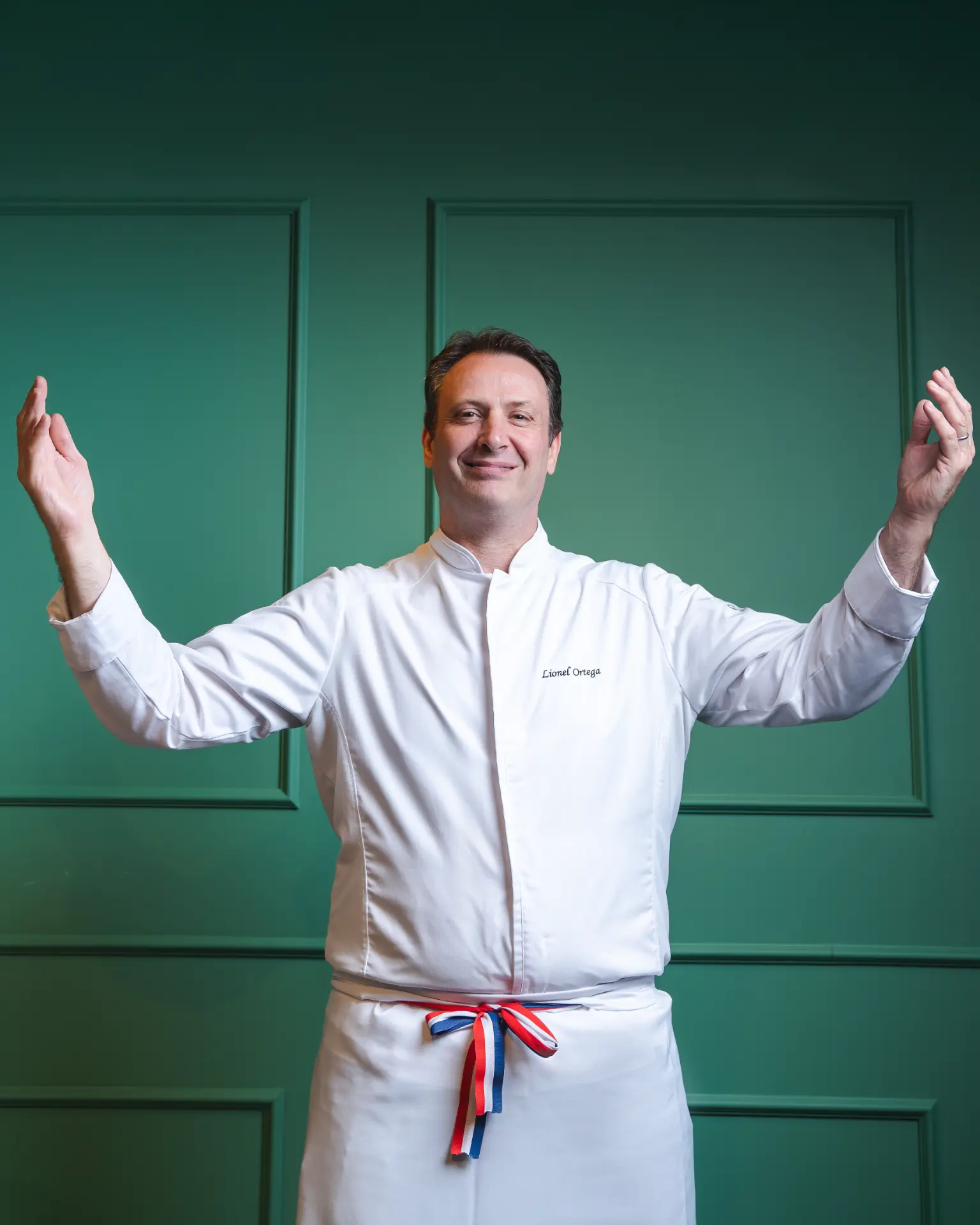 Chef Lionel Ortega de dólmã branca e avental com fita azul, branca e vermelha, com os braços abertos diante de uma parede verde.